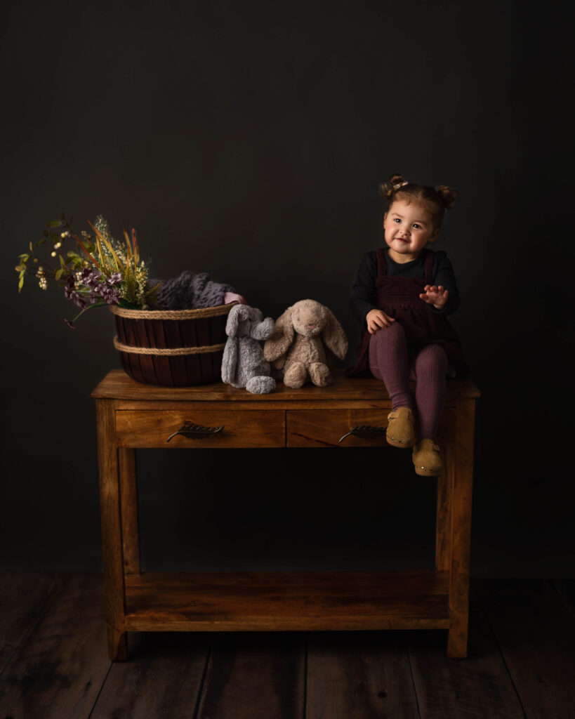 Little girl waiting for the arrival of her baby brother sitting on a wooden table with flowers in a basket with jellycat teddys - Little Blue Boutique Calabasas