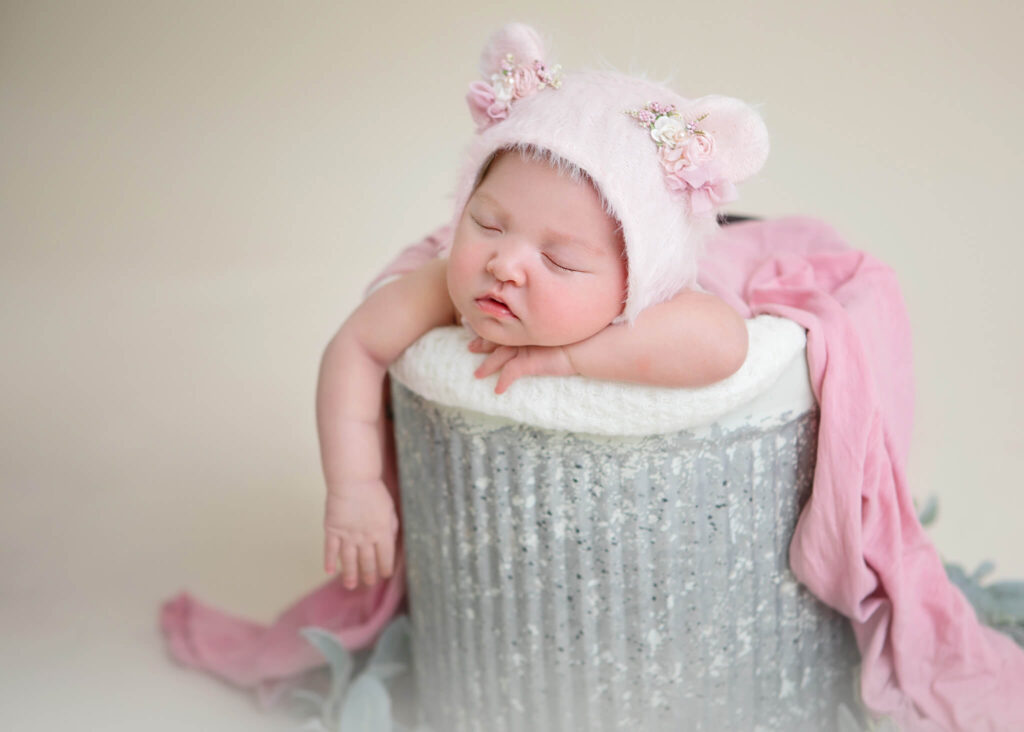Image of a newborn in a bucket with a pink teddy bear hat on. 