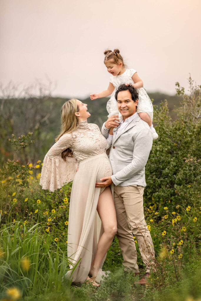 Family laughing at maternity photoshoot in Los Angeles with Elsie Rose Photography. Mom wearing beige dress, dad with grey blazer and daughter in white lace dress