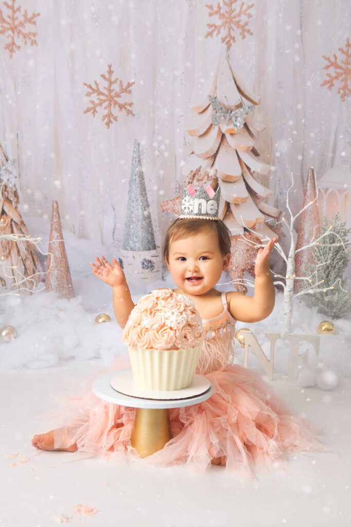 Happy baby girl smiling wearing a pink dress and sitting behind a large pink cupcake for her birthday photoshoot - Best Birthday Cakes in Los Angeles