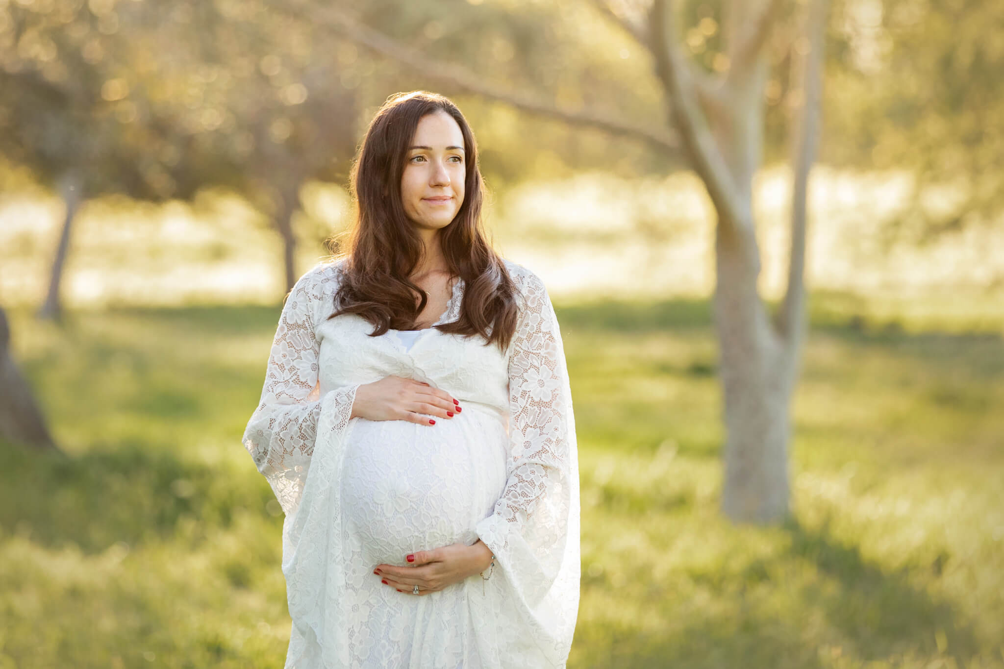Mom to be holding her baby bump and wearing a white lace dress looking into the distance - Hatch Brentwood