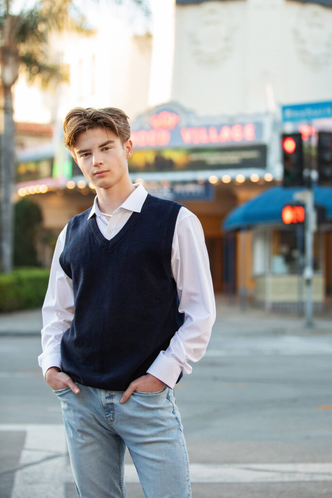 Boy wearing white shirt, blue sweater and jeans in front of movie theatre in Westwood - High School Senior Portraits