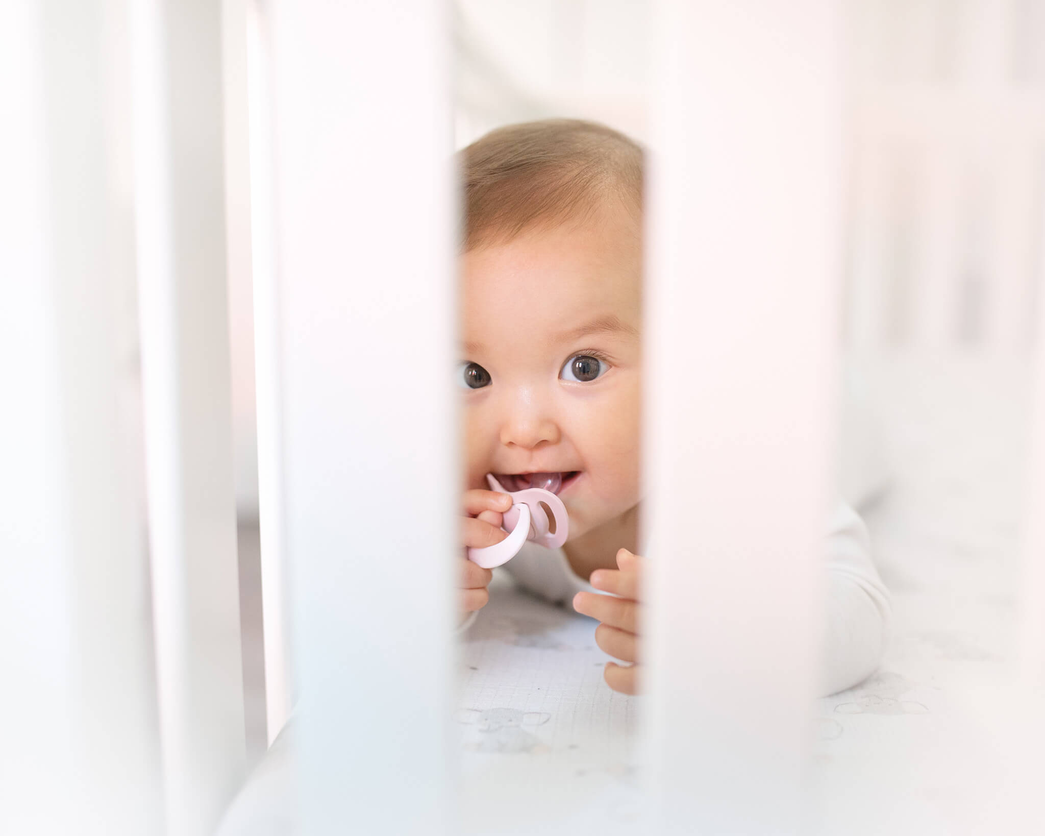 Baby in a crib with a pacifier smiling. All white image taken by Elsie Rose Photography - Los Angeles Lactation Consultant