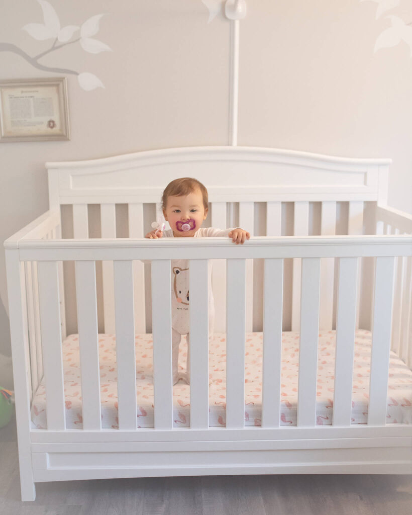 Baby standing in crib with a pacifier, image white with touches of pink - baby sleep consultant los angeles