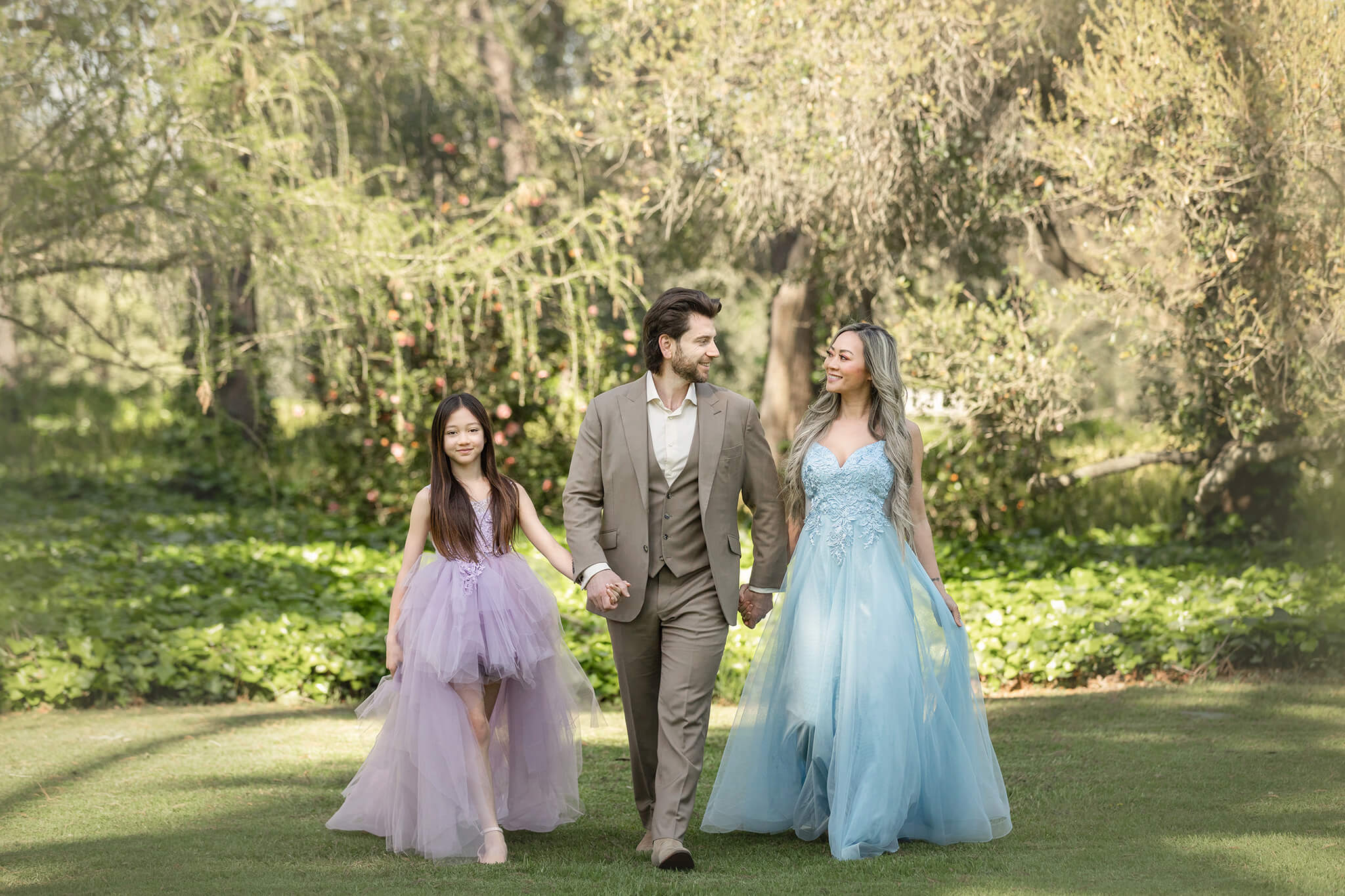 Family photo of mom, dad and daughter at Los Angeles Arboretum. Mom in a blue dress, dad in a suit and daughter in a purple dress walking towards camera.