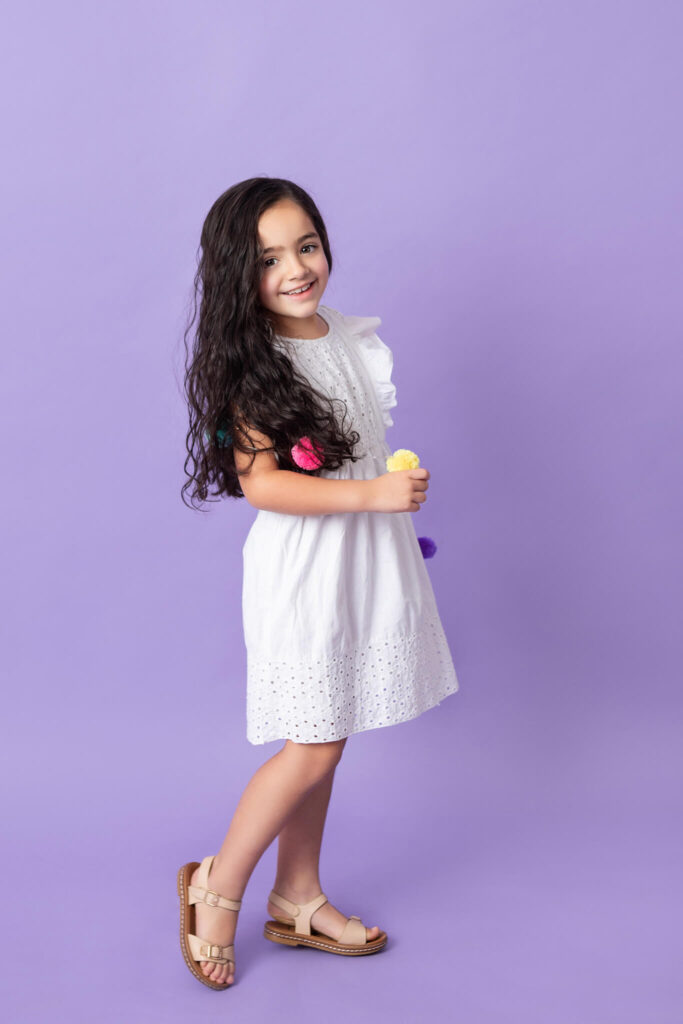 Little girl on purple backdrop photographed by Elsie Rose Photography. Wearing white dress.