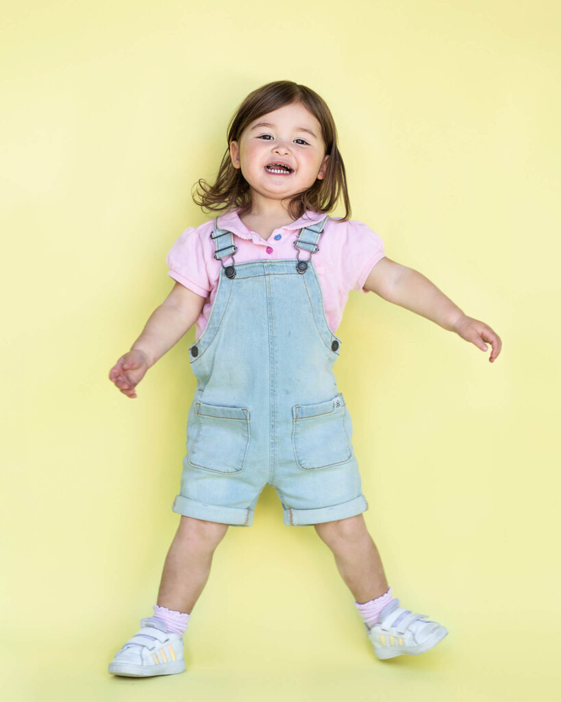 Toddler on yellow backdrop wearing dungarees and laughing - pediatric dentist los angeles