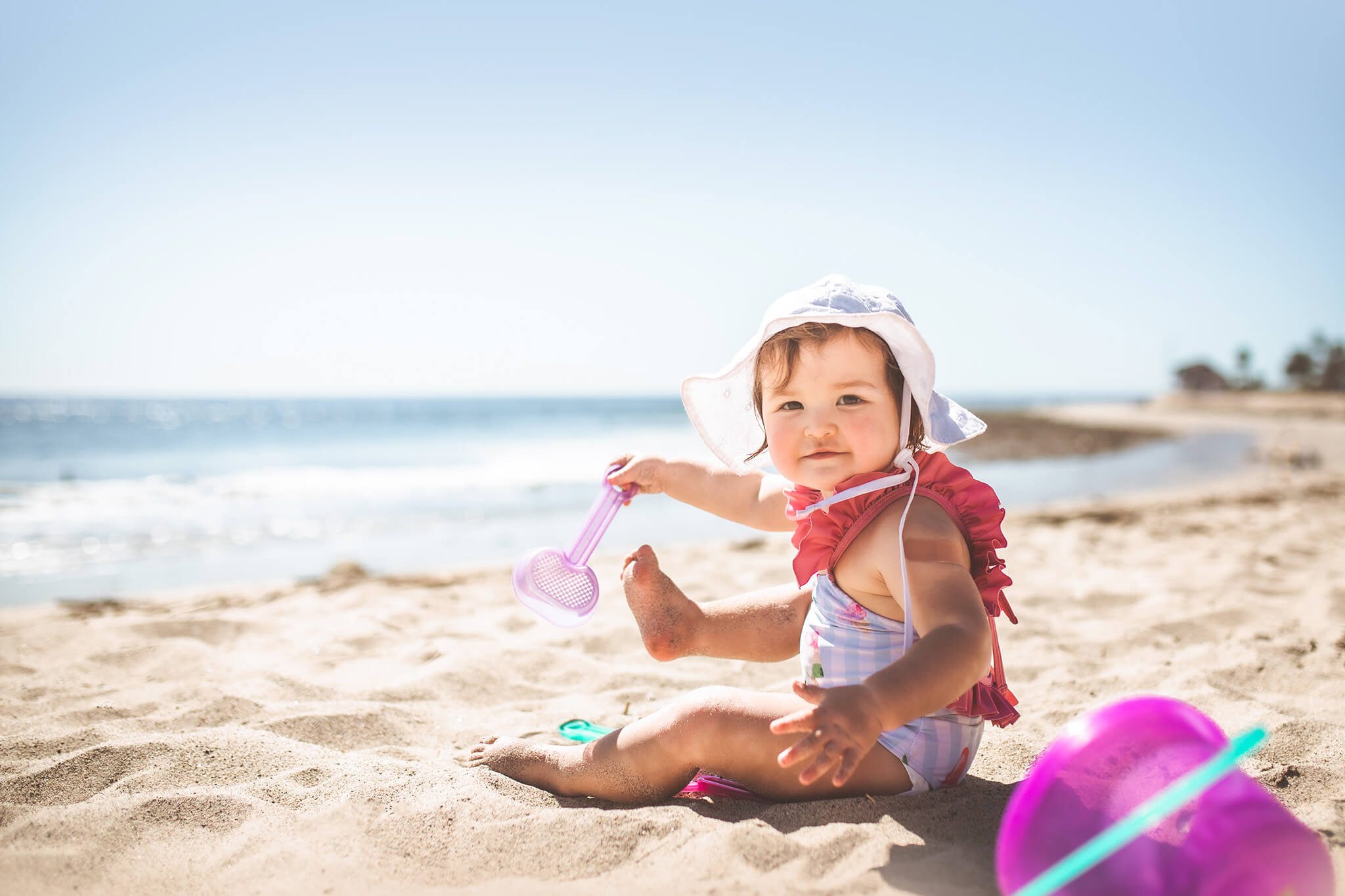 Baby girl sitting on the beach in Malibu with her spade and bucket, wearing a white hat and swimsuit - swim lessons for toddlers los angeles
