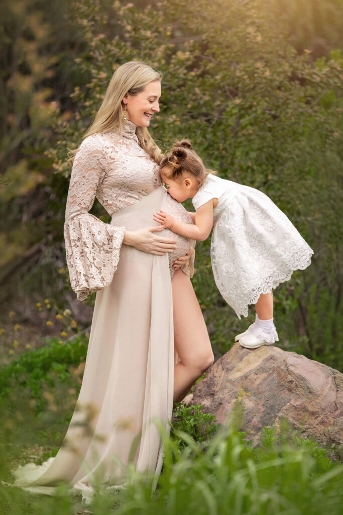 Little girl standing on a rock and kissing moms baby bum. Little girl wearing white and mom in beige. - Women's Care of Beverly Hills.