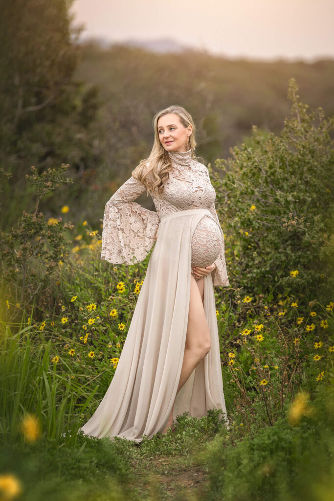 Mom to be surrounded by flowers in the park with sunset behind in a maternity photoshoot wearing a beige lace dress.