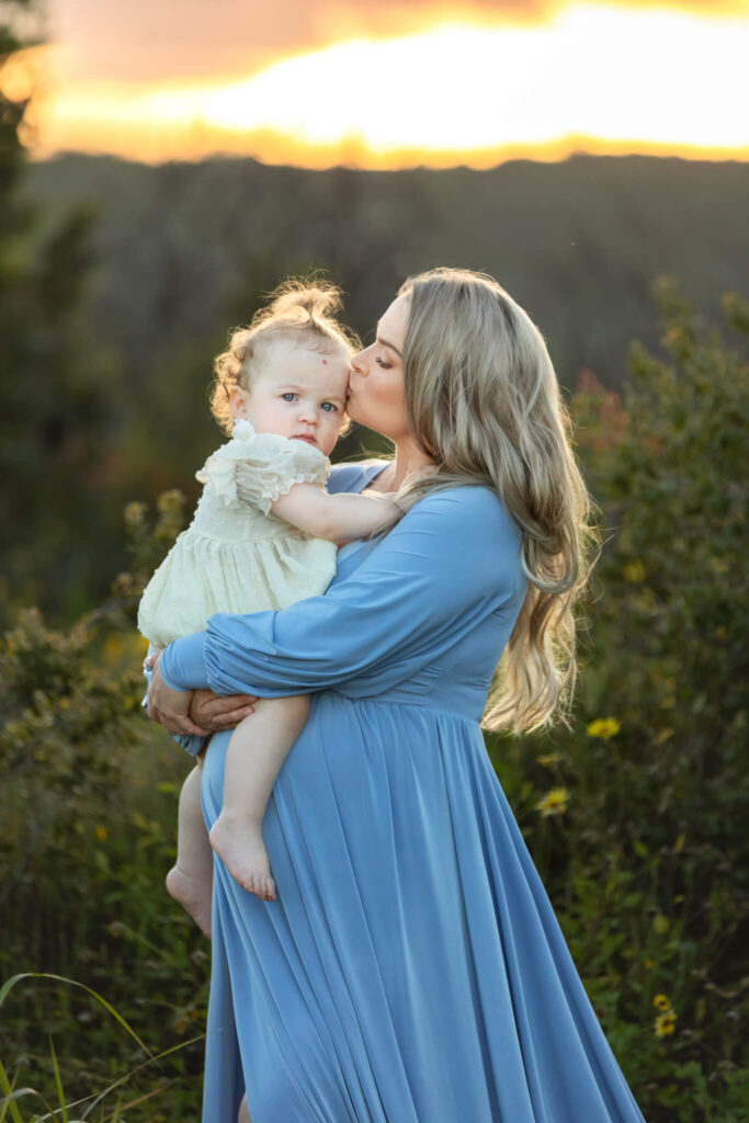Mom with her toddler daughter, pregnant with second baby. Wearing a blue dress with sunset behind