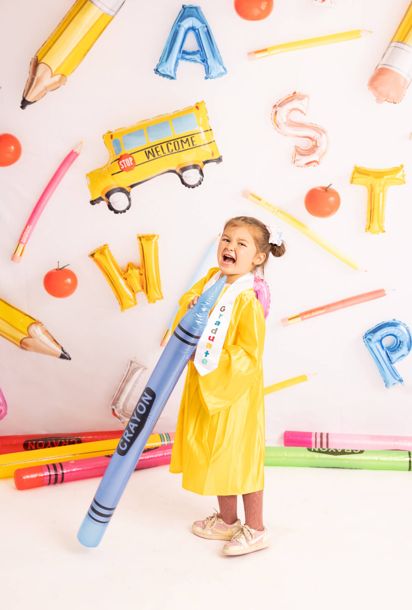 Little girl in yellow gown graduating preschool and laughing with a big decorative blue crayon